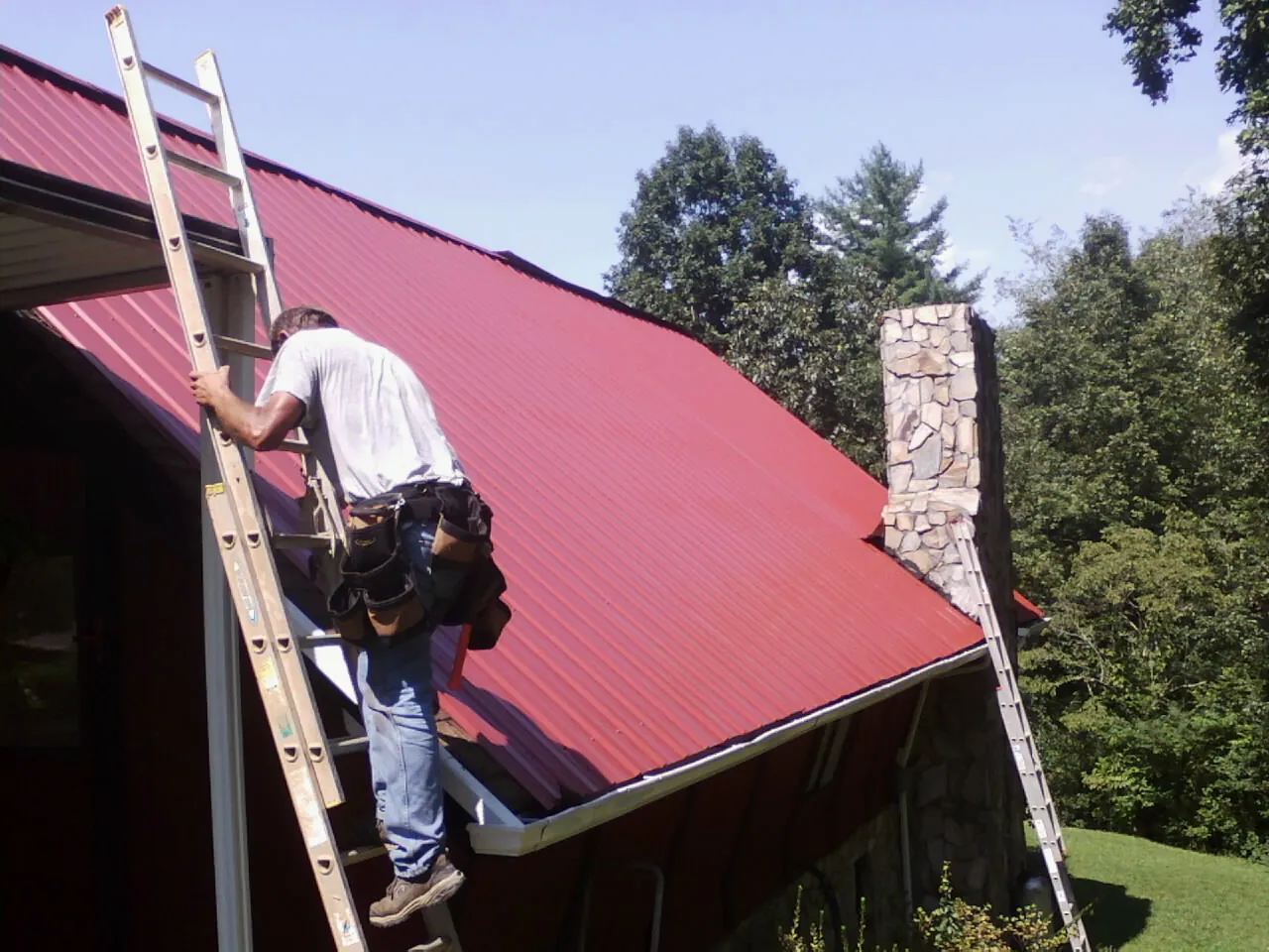 Roofer on a ladder beside a red metal roof and stone chimney — Soffit & Fascia Repair by Roofing Brothers in Milligan